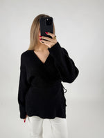 Person tager et billede med en smartphone, iført en sort sweater og hvide bukser. Baggrunden er enkel og hvid, hvilket fremhæver tøjet og personen tydeligt.