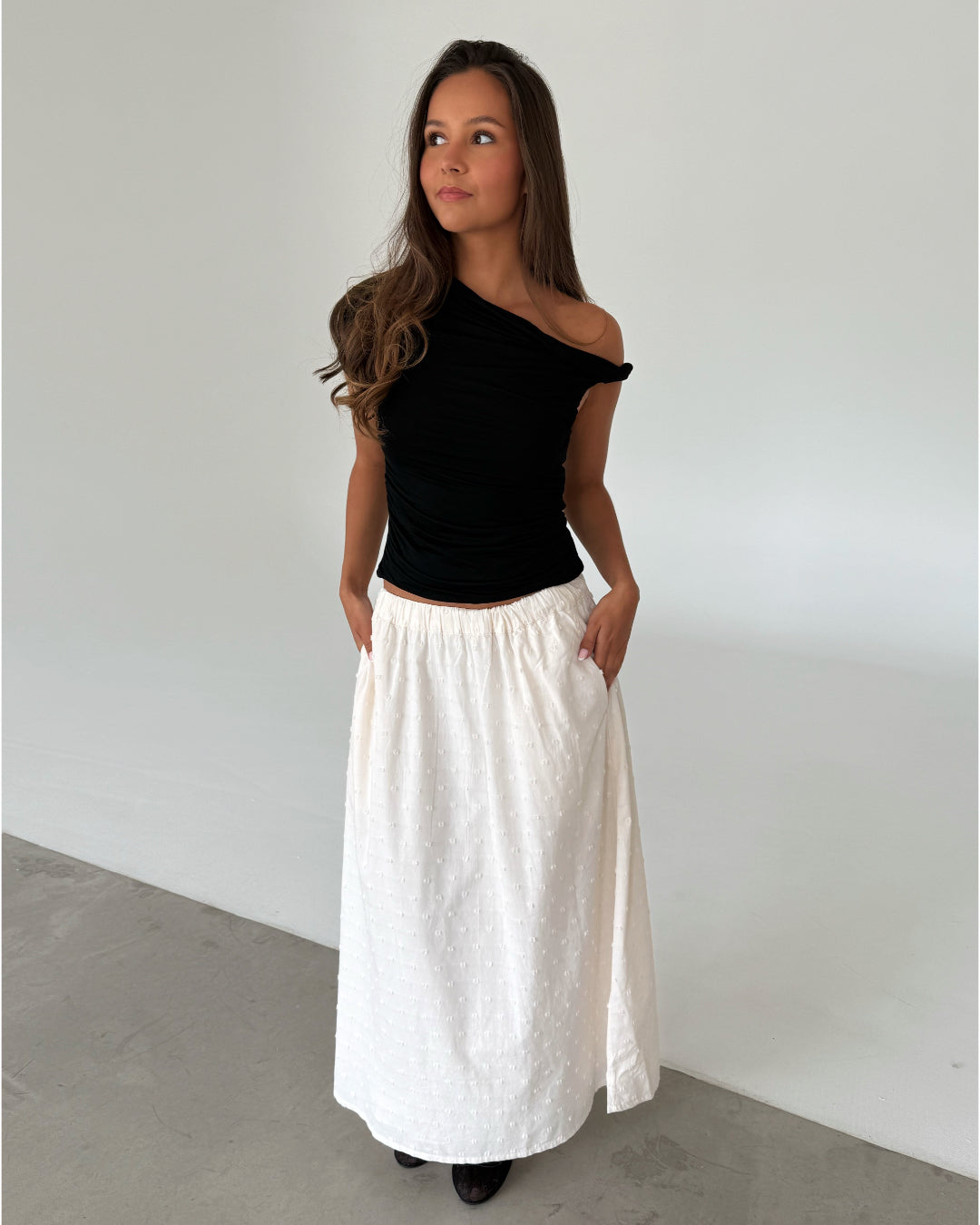Modelfoto af pige som bærer en sort oneshoulder MILENA top, samt en hvid nederdel og sorte ballerinasko. Alt er fra BySofieSønderby