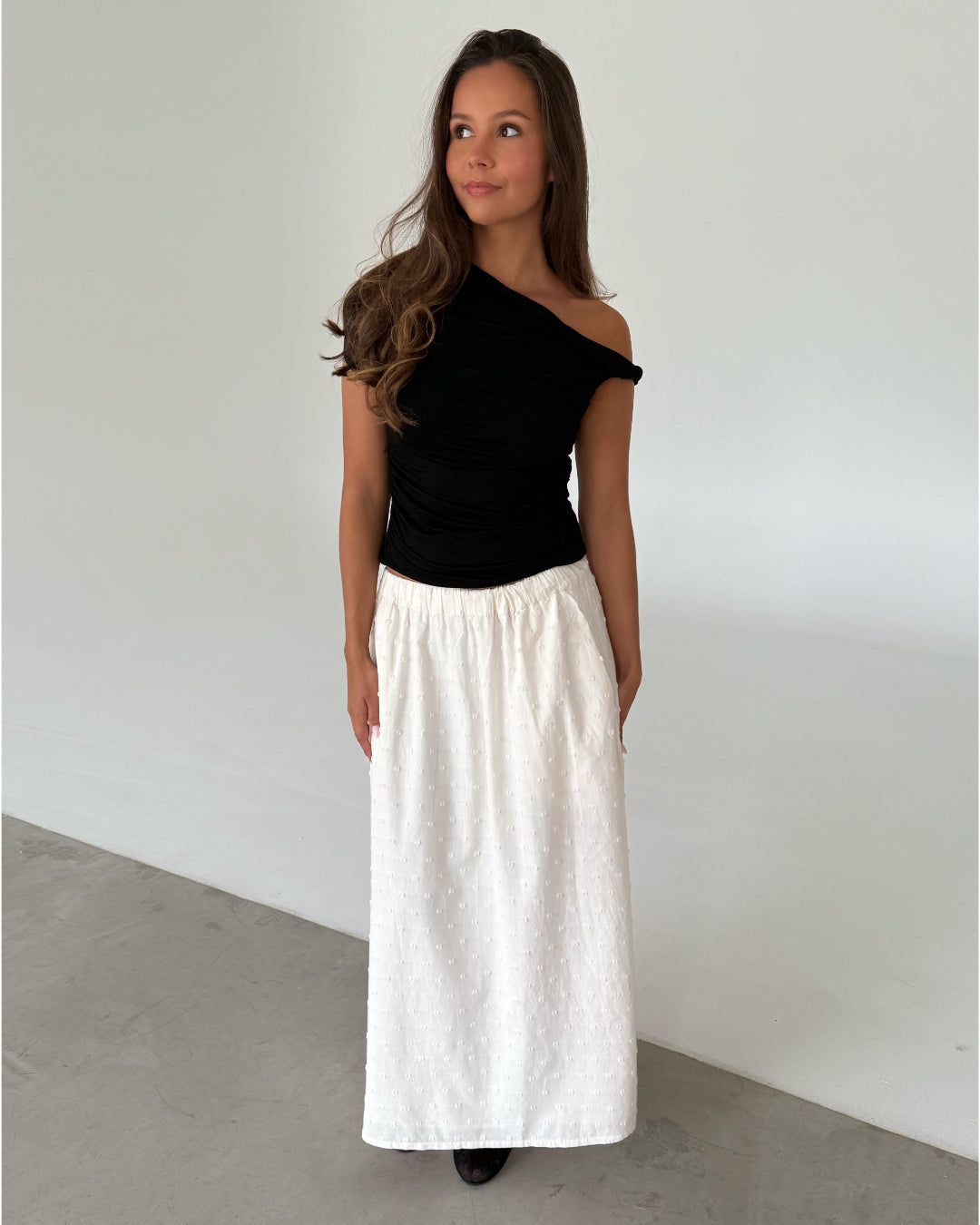Modelfoto af pige som bærer en sort oneshoulder MILENA top, samt en hvid nederdel og sorte ballerinasko. Alt er fra BySofieSønderby