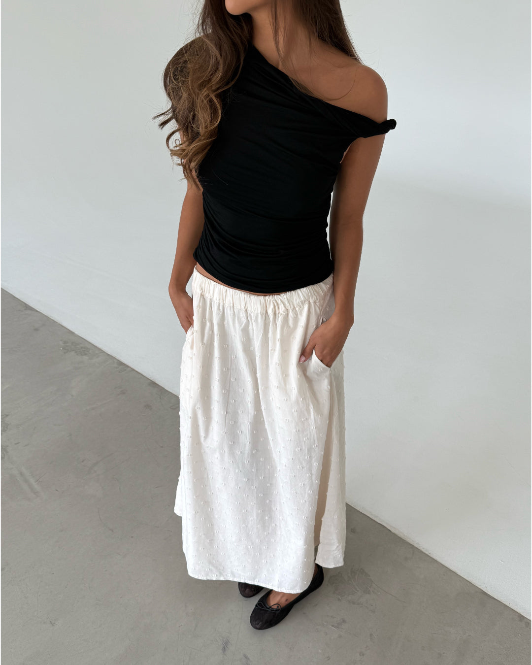 Modelfoto af pige som bærer en sort oneshoulder MILENA top, samt en hvid nederdel og sorte ballerinasko. Alt er fra BySofieSønderby