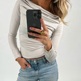 En person med hvid bluse, blå jeans og smykker holder en sort smartphone, mens vedkommende tager et billede mod en neutral baggrund.