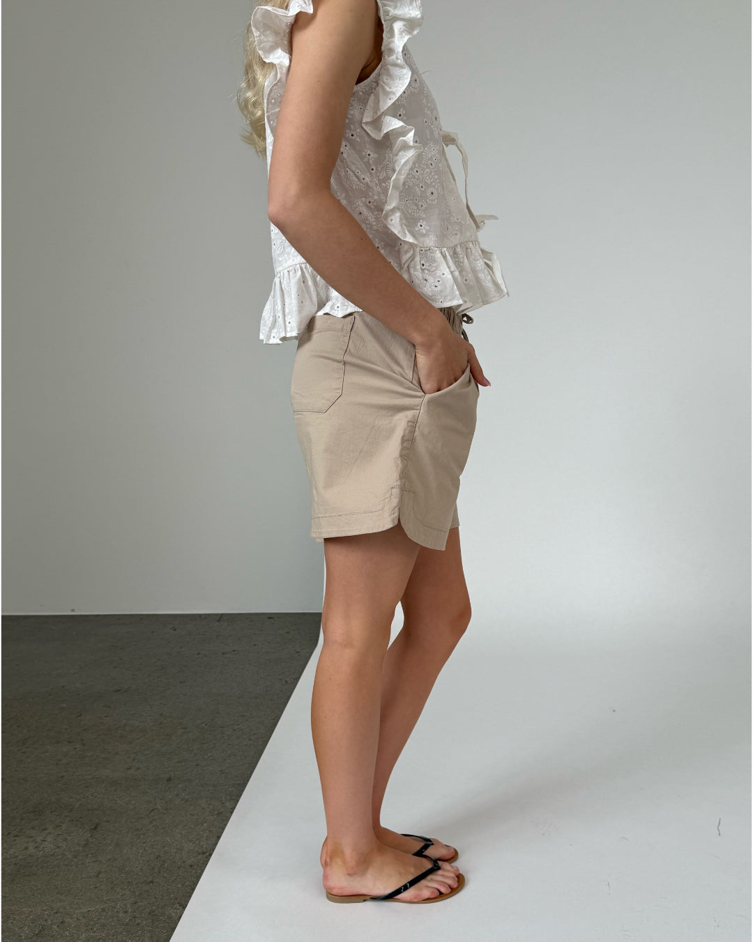 En person står i profil iført en hvid top med flæser og beige shorts, hvor hænderne hviler i lommerne, på et gulv med grå og hvid baggrund.