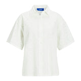 Hvid camisola med korte ærmer og hulmønster. Den har en klassisk krave og knaplukning, hænger frit mod en simpel, hvid baggrund. Etiket med teksten "JJXX" indeni.