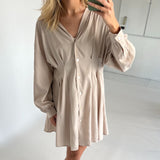 En person iført en beige kjole tager en selfie med en smartphone i et lyst rum. Kjolen har knapper foran og brede ærmer.