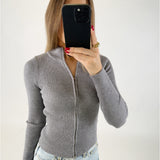 En person holder en mobiltelefon foran ansigtet, iført en grå striktrøje med lynlås og lyseblå jeans. Baggrunden er hvid og enkel.