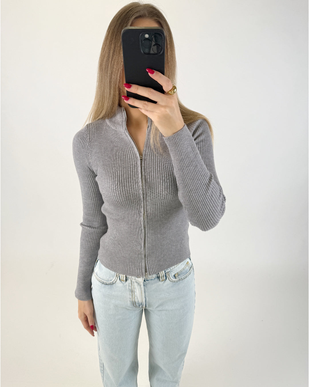 Kvinde i grå, ribstrikket trøje og lyse jeans tager selfie med sort mobiltelefon foran ansigtet i et lyst rum. Rød neglelak og guld ring ses.