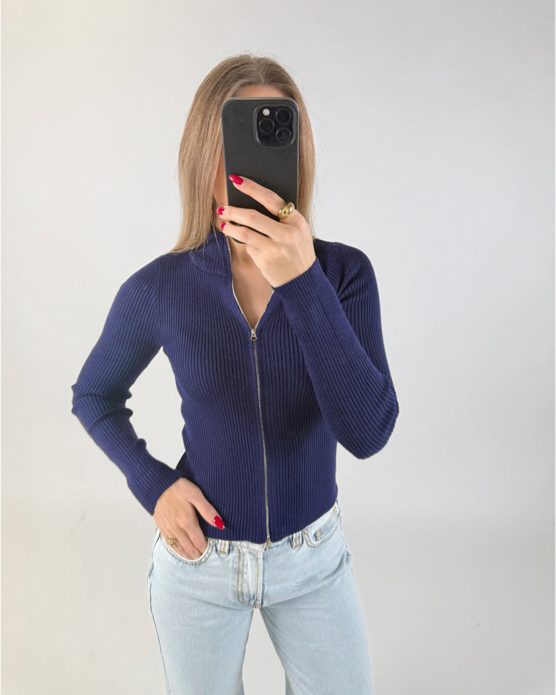Kvinde med langt hår holder en sort smartphone op foran ansigtet. Hun har en mørkeblå trøje med lynlås og lyse jeans. Baggrunden er en simpel, lys væg.