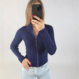 Kvinde med langt hår holder en sort smartphone op foran ansigtet. Hun har en mørkeblå trøje med lynlås og lyse jeans. Baggrunden er en simpel, lys væg.