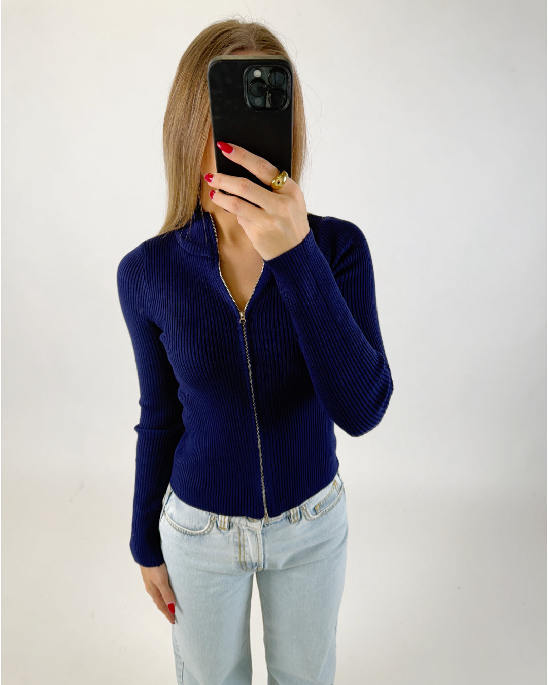 En person med langt hår holder en smartphone foran ansigtet i en selfie-positur. De har en mørkeblå trøje med lynlås og lyse jeans på, i et neutralt rum.