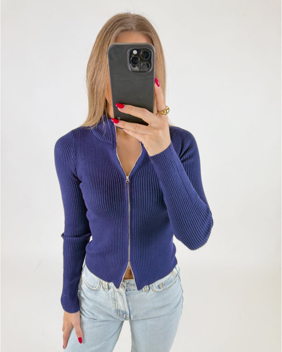 Kvinde holder en sort telefon foran ansigtet, iført en mørkeblå, ribbet trøje med lynlås, kombineret med lyse jeans. Baggrunden er hvid og minimalistisk.