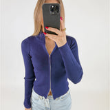 Kvinde holder en sort telefon foran ansigtet, iført en mørkeblå, ribbet trøje med lynlås, kombineret med lyse jeans. Baggrunden er hvid og minimalistisk.