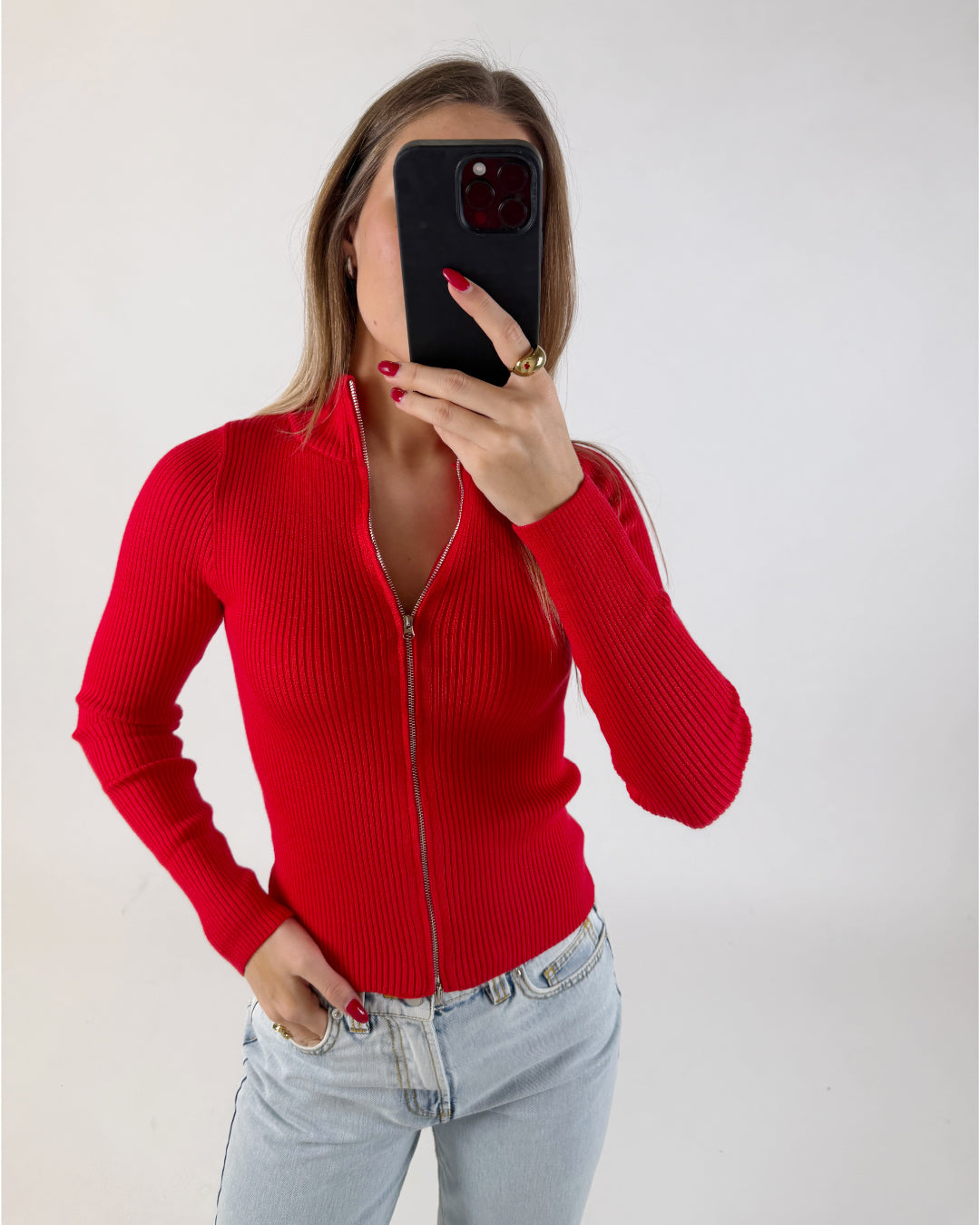 Person iført rød striktrøje tager selfie med sort telefon i lyseblå jeans mod neutral, lys baggrund.
