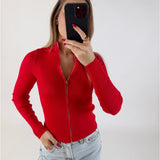 Person iført rød striktrøje tager selfie med sort telefon i lyseblå jeans mod neutral, lys baggrund.