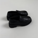 Et par sorte loafers står side om side på en lys baggrund. Skoene har en robust, rillet sål og en glat overflade med en strop over forfoden.
