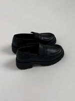 Et par sorte loafers står side om side på en lys baggrund. Skoene har en robust, rillet sål og en glat overflade med en strop over forfoden.