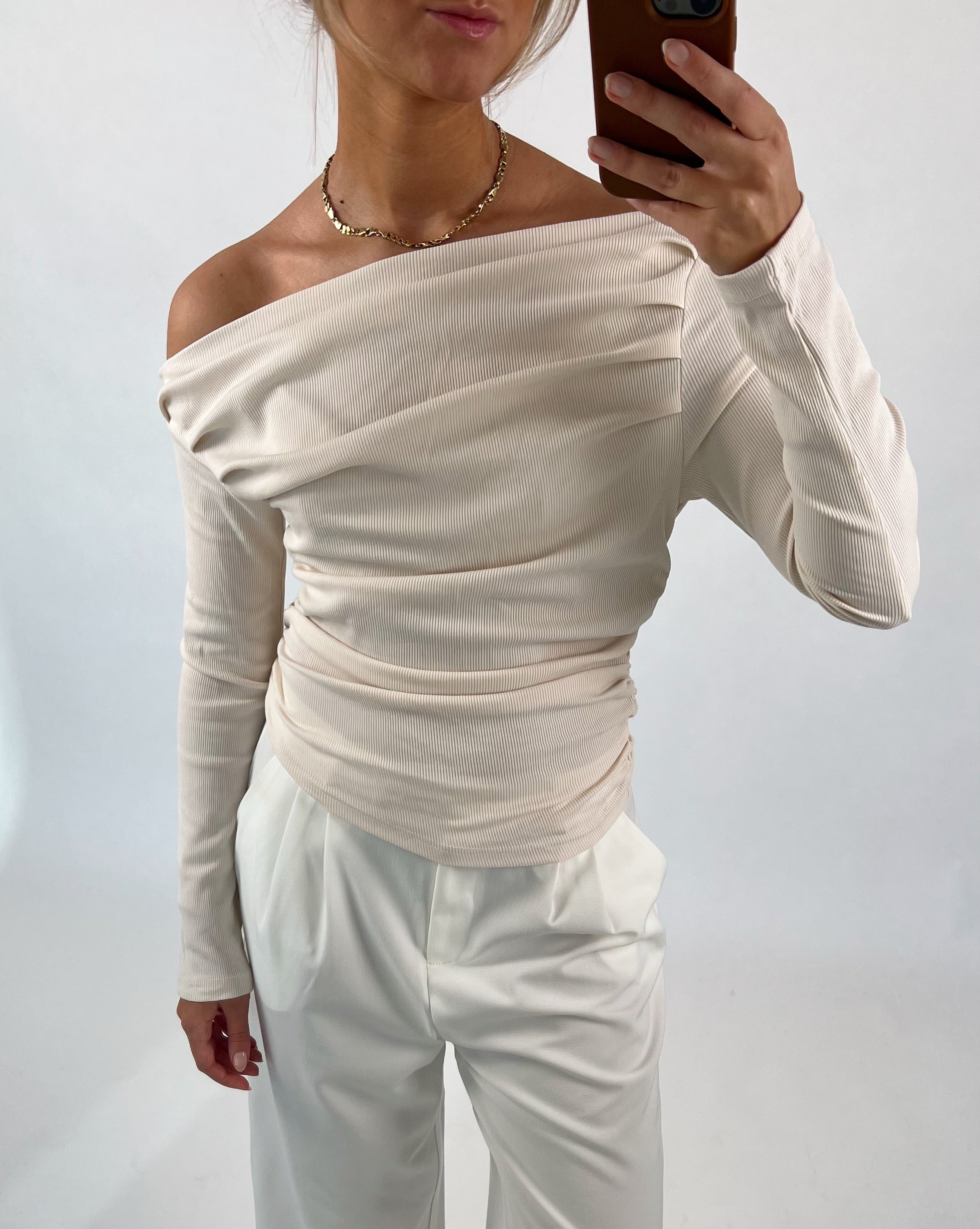 En person tager et selfie iført en off-shoulder cremefarvet top og hvide bukser, stående i et neutralt, ensfarvet rum.