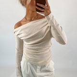 Person tager selfie iført cremefarvet off-shoulder bluse og hvide bukser, står i en neutral, lys baggrund.
