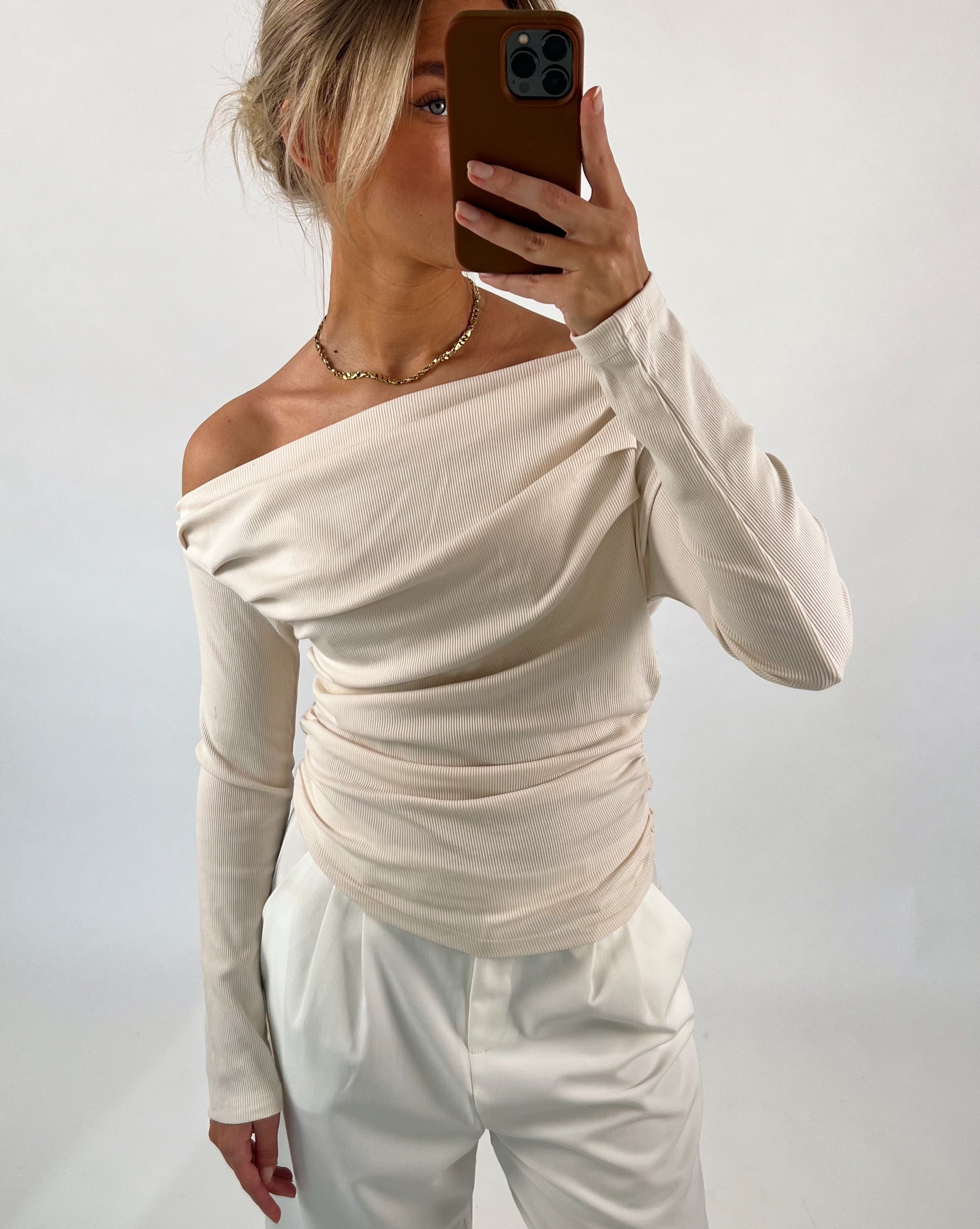 Person tager selfie iført cremefarvet off-shoulder bluse og hvide bukser, står i en neutral, lys baggrund.
