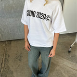 En person står iført en hvid t-shirt med teksten "STUDIO 2020©," grønne bukser og sandaler, på et betonunderlag foran en grå væg.