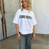 En person med blondt, krøllet hår står, iført en hvid T-shirt med teksten "STUDIO 2020©" og jeans. Baggrunden er rustikt med metaldetaljer og en minimalistisk væg.