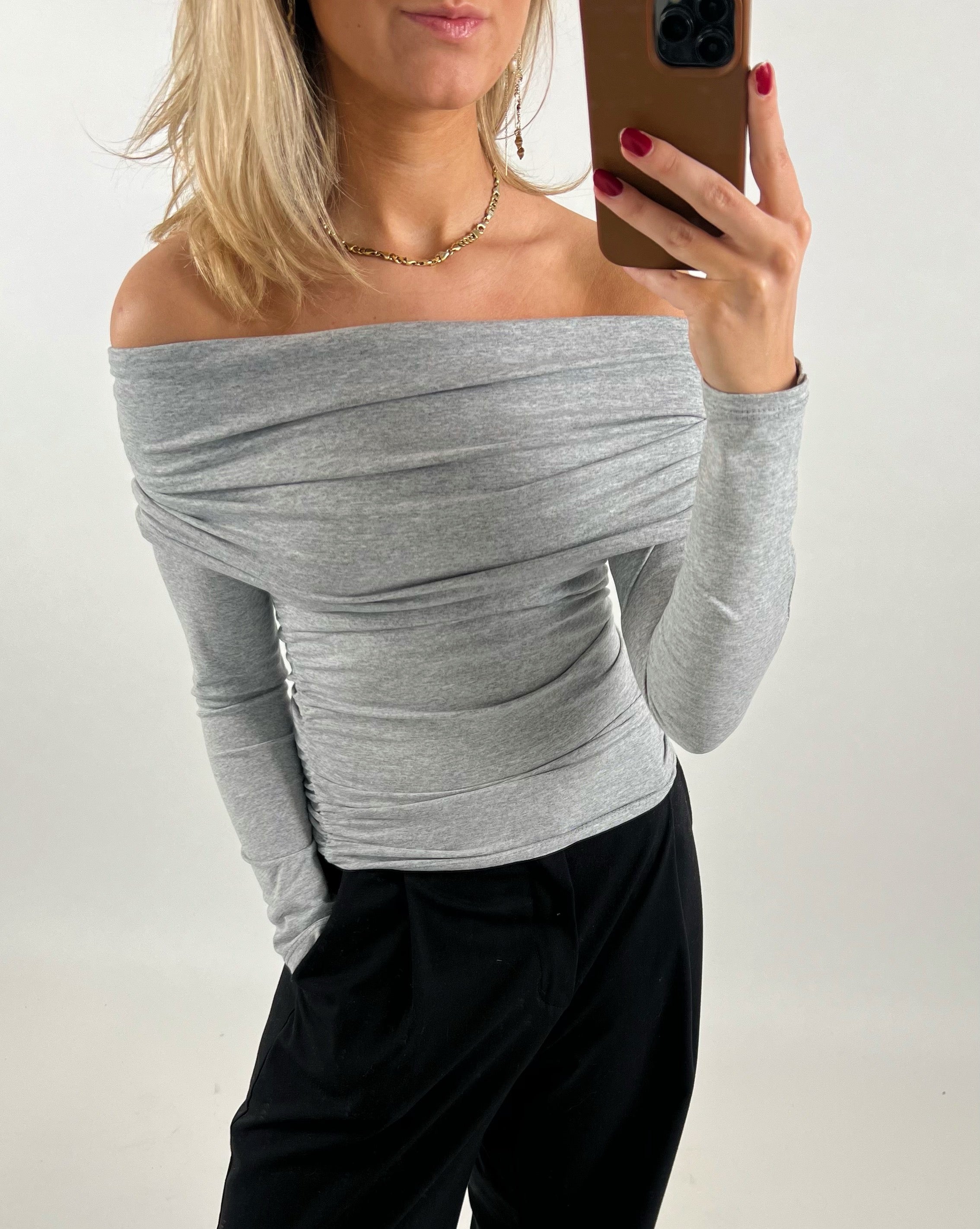 En person iført en grå off-shoulder bluse tager et spejl-selfie med en smartphone. Personen bærer sorte bukser og står mod en hvid baggrund.