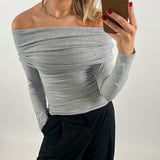 En person iført en grå off-shoulder bluse, tager en selfie med en smartphone, mens hun står i et rum med lys baggrund. Hun har rød neglelak og bærer sort bukser.