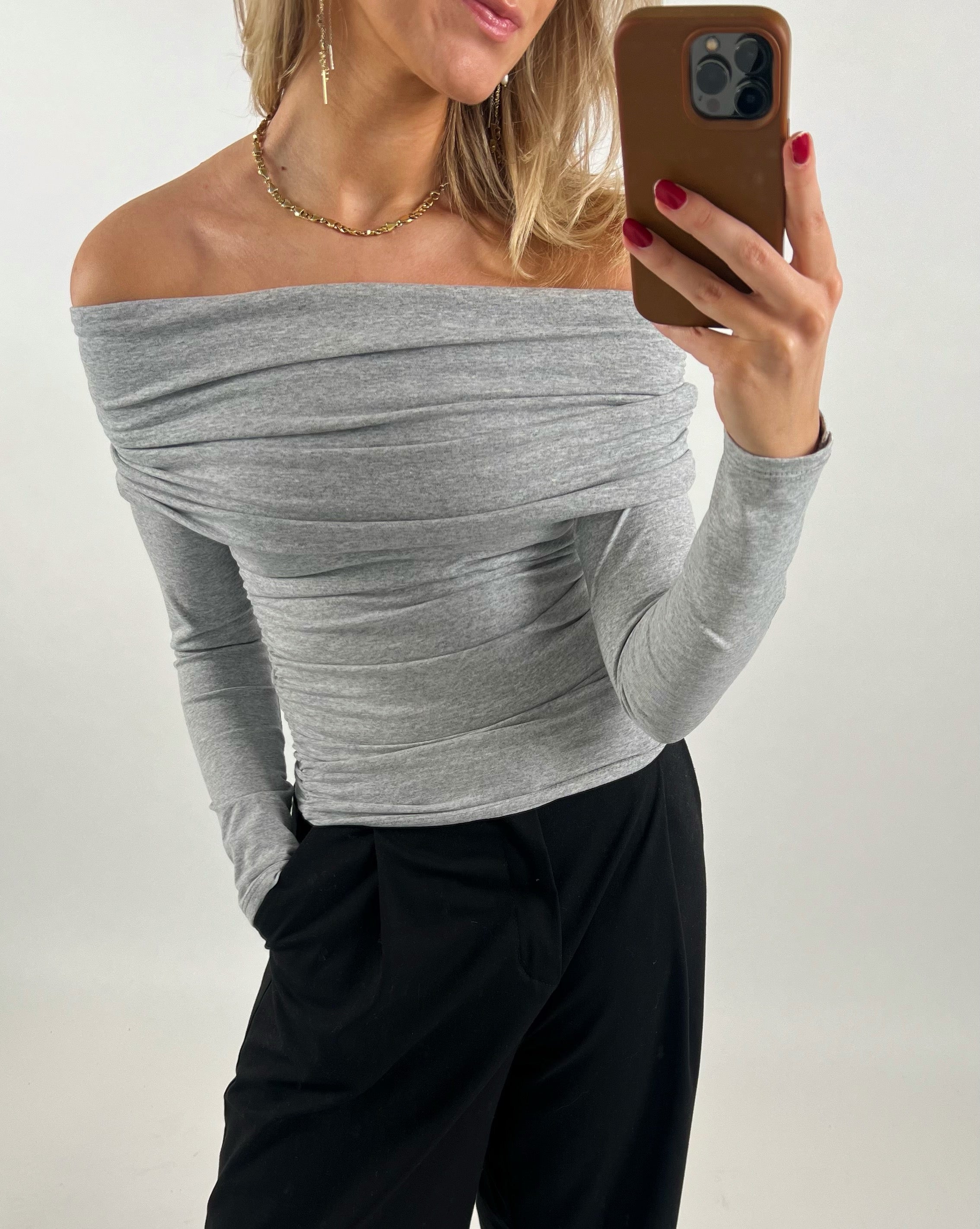En person iført en grå off-shoulder bluse, tager en selfie med en smartphone, mens hun står i et rum med lys baggrund. Hun har rød neglelak og bærer sort bukser.