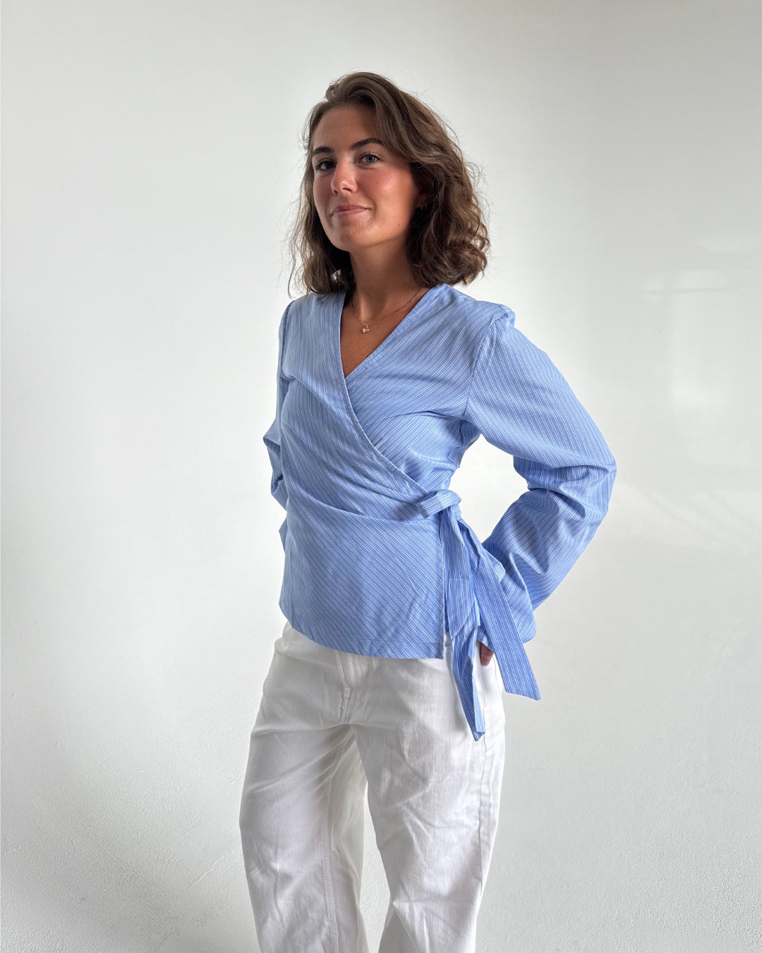 Modelfoto af pige som bærer en slå-om skjorte i lyseblå med striber, samt et par hvide Nanna jeans. Baggrund hvid. Alt fra By Sofie Sønderby