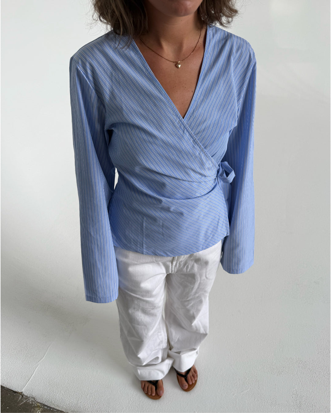 Modelfoto af pige som bærer en slå-om skjorte i lyseblå med striber, samt et par hvide Nanna jeans og Carmen sandaler. Baggrund hvid. Alt fra By Sofie Sønderby