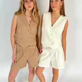 To kvinder iført matchende vest-og-shorts-sæt, den ene i beige, den anden i hvid, poserer sammen mod en hvid baggrund, iført afslappet fodtøj og smykker.