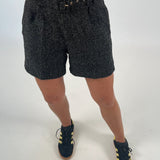 En person iført sorte shorts med mønster står med en hånd ved taljen. De bærer også sorte og gule sneakers på en hvid baggrund.