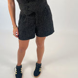 Person iført sort mønstret shorts og top, står på et hvidt gulv. De bærer sorte sneakers med gule striber og guldsmykker på håndleddet.