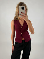 En person holder en mobiltelefon op foran ansigtet, iført en ærmeløs, rød top og sorte bukser, står i et neutralt belyst rum.