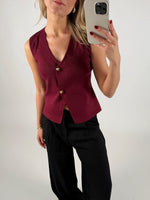 En person iført en vinrød vest med guldfarvede knapper tager et spejlbillede med en smartphone. De har sorte bukser på, og baggrunden er lys grå.