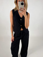 En person i sort tøj tager et spejlbillede med en smartphone i et neutralt, lyst rum. Personen bærer en sort vest og bukser og har rød neglelak.