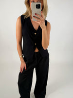 En person iført sort vest og bukser tager et spejlbillede med en smartphone i en lys baggrund. Personen har rødlakerede negle og en gylden armbånd.