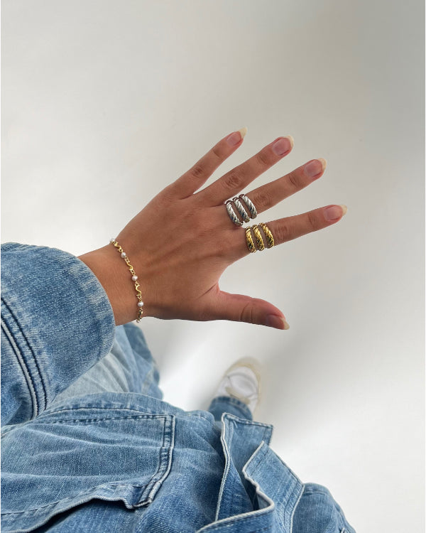 En hånd bærer to flettede ringe og et armbånd. Personen iført blå denimjakke og jeans står på en lys baggrund.