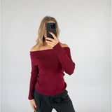 En person iført en mørkerød off-shoulder trøje tager et spejlbillede med en sort telefon i et minimalistisk rum med lysegrå baggrund.