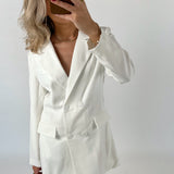 En person iført en hvid blazer-kjole tager et selfie med en brun telefon, mens de står foran en enkel, hvid baggrund.