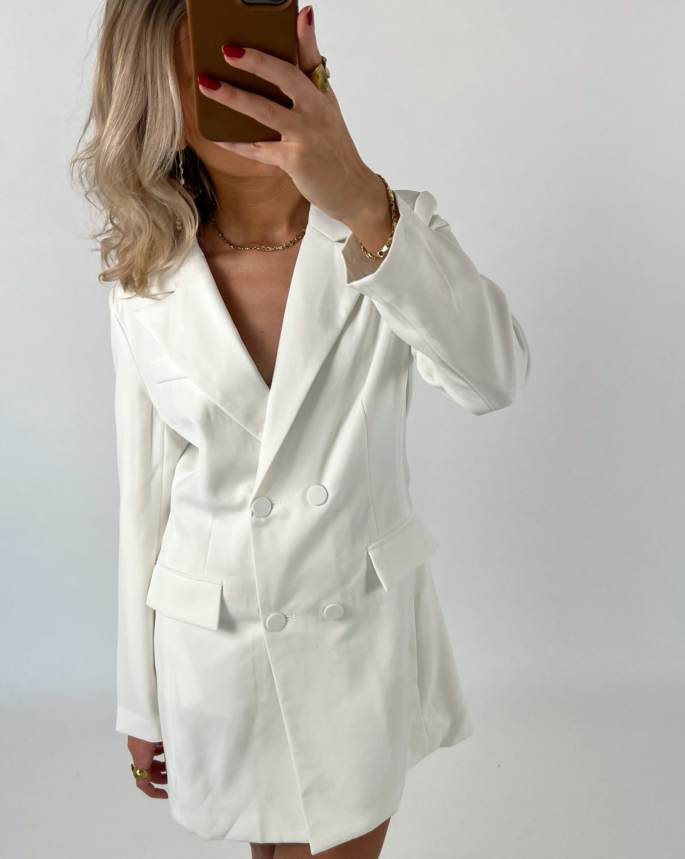 En person iført en hvid blazer-kjole tager et selfie med en brun telefon, mens de står foran en enkel, hvid baggrund.