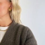 En person med lyst hår bærer en guldfarvet blomsterørering. Personen er iført en brun striktrøje og en hvid bluse med halskæde, mod en neutral baggrund.