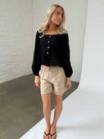 En kvinde iført sort bluse og beige shorts står afslappet, kigger til siden. Baggrunden er en væg af hvide mursten og et gråt gulv i et minimalistisk rum.