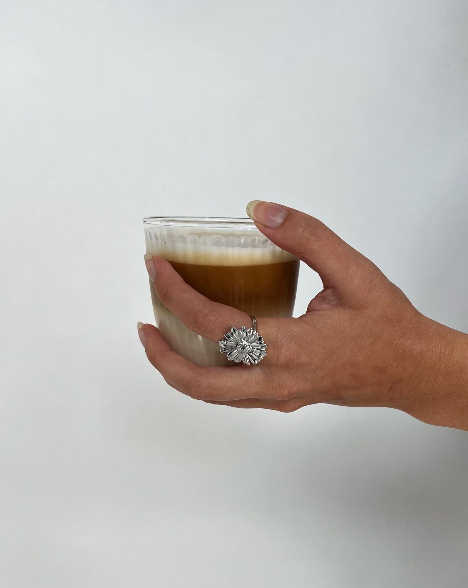 Hånden holder forsigtigt et glas med lagdelt kaffe og mælk. En dekorativ ring med en blomst er synlig på fingeren. Baggrunden er simpel og lys.