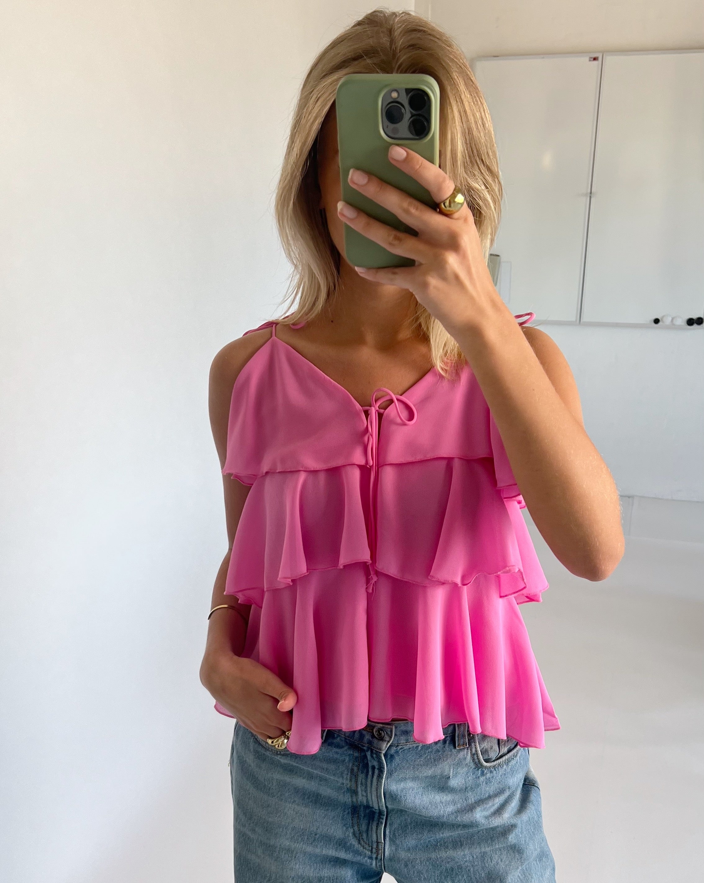Person tager et spejlbillede med en grøn telefon, iført en lyserød top med flæser og lyse jeans. Bagved er der en hvid væg og et whiteboard.