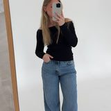 En person i sort trøje og blå jeans tager et spejlbillede med en telefon. Personen står i et lyst rum med en hvid væg og trægulv.
