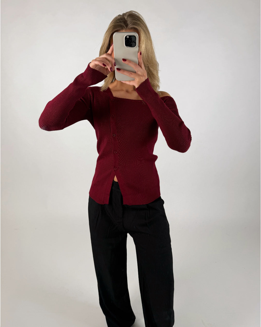 Person tager billede af sig selv med smartphone, iført bordeaux cardigan og sorte bukser, står i et neutralt, lysegråt rum.