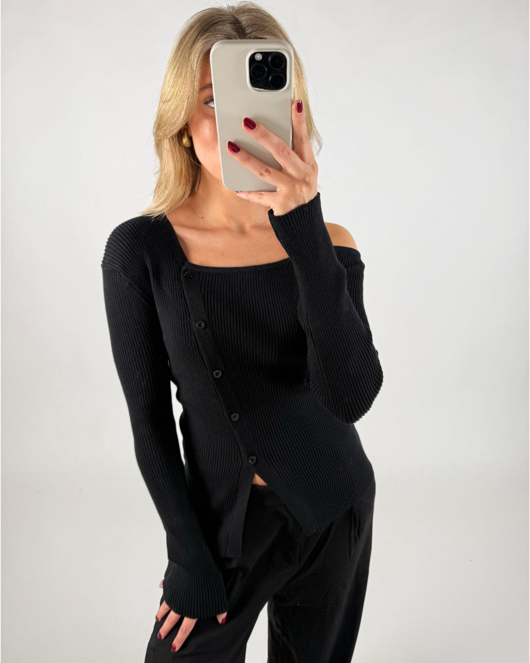 En person i sort trøje tager et selfie med en smartphone i et minimalistisk rum. Hendes hår er lyst, og hun har rød neglelak.