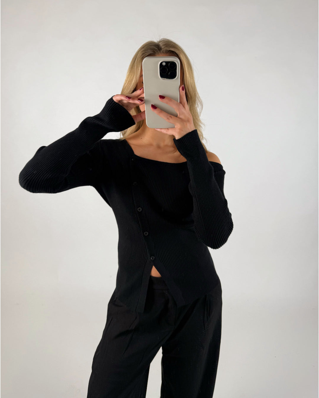 En person i sort tøj tager et spejlselfie med en mobiltelefon. Personen står i et enkelt, lyst rum uden synlige møbler.