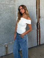 En ung kvinde står afslappet med hånden i lommen. Hun har en hvid off-shoulder-top og jeans på. Baggrunden er en rustik metalvæg.