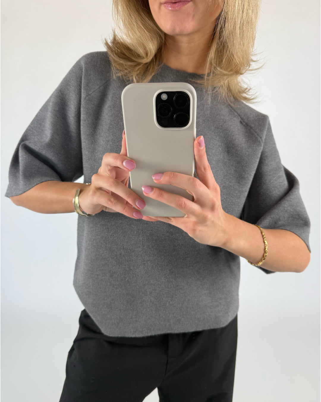 En person iført en grå bluse tager et billede med en smartphone. Personen står i et indendørs, neutralt oplyst rum og bærer et armbånd.